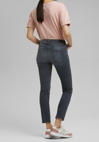Mujer con cabello oscuro y largo, vistiendo una camiseta rosa de manga corta, jeans de corte slim en gris oscuro y zapatillas blancas con detalles rosas, de pie de espaldas.
