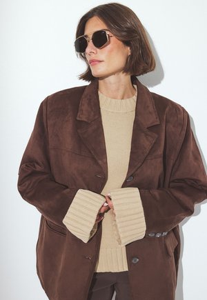 Woman with short brown hair wears large dark sunglasses, oversized brown suede coat, and beige knit sweater with extended cuffs.