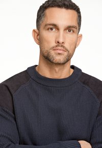 Dunkelblauer Pullover mit gerippter Struktur und glatten Wildlederakzenten an den Schultern. Rundhalsausschnitt und entspannte Passform.