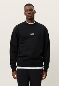 Άνδρας που φοράει μαύρο φούτερ και παντελόνι με μικρές ορθογώνιες ετικέτες λογότυπου "LES DEUX" στο στήθος και στην αριστερή μηρό, μπροστά σε απλό φόντο.