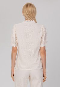 Blouse blanche en broderie anglaise avec des manches bouffantes, un col volanté et des accents de boutons. Présente un motif texturé de découpes circulaires.