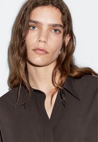 Tmavě hnědá košile s límcem na knoflíky, vyrobená z hladkého materiálu s jemným prošíváním. Model má dlouhé vlnité vlasy.