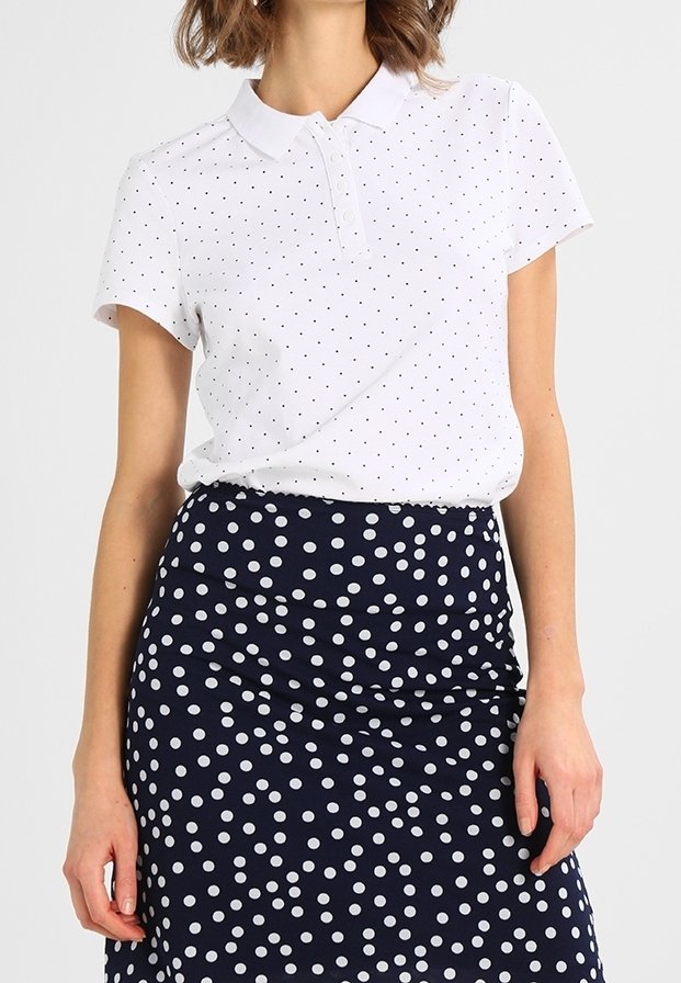 Femme portant un polo blanc à manches courtes avec de petits pois noirs et une jupe taille haute bleu marine à gros pois blancs.