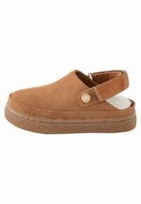Chaussure slip-on en suède marron avec une semelle en caoutchouc texturée, une sangle arrière ajustable et une doublure douce visible au niveau du talon.
