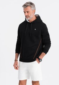 Sudadera con capucha negra y detalles en naranja, hecha de una tela suave. Cuenta con un bolsillo tipo canguro en la parte delantera y puños acanalados.