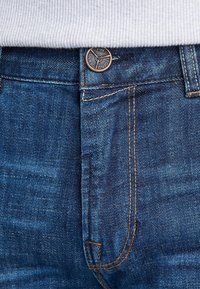 Jeans de mezclilla azul oscuro con un corte clásico, que presentan un acento de botón en tono cobre, costuras visibles y un detalle en el bolsillo delantero.