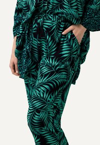 Grön bladmönstrad tyg i en avslappnad passform som har dragsko i midjan och sidoficka, placerad mot en mörk bakgrund.