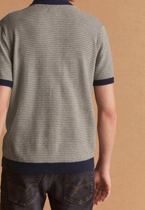 Polo à manches courtes de couleur gris clair avec un motif texturé, col et ourlet bleu marine. Porté avec un jean foncé montrant les détails de la poche arrière.