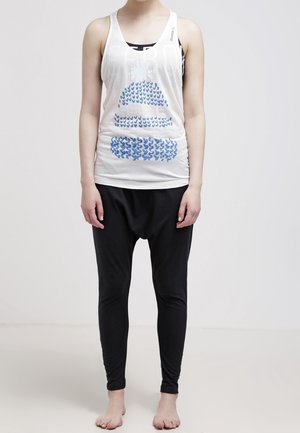 Personne portant un débardeur blanc sans manches avec un motif bleu et un pantalon noir ample, debout pieds nus devant un fond uni.