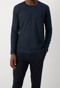 Granatowa koszulka z długim rękawem wykonana z miękkiej bawełny, z okrągłym dekoltem i subtelnym logo na klatce piersiowej, zestawiona z ciemnymi spodniami.
