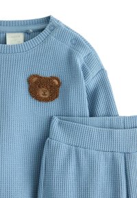 Lysblåt waffle-strik to-dels sæt med bjørne-applikation på toppen. Har tryknapper på skulderen og ribbede manchetter på bukserne.