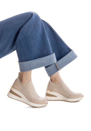 Zapatillas de punto beige con tacón cuña, que presentan detalles en dorado y una superficie texturizada, combinadas con pantalones de mezclilla enrollados.