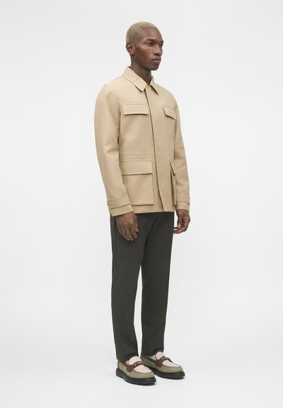 Άνδρας με κοντά ξανθά μαλλιά που φοράει μπεζ μπουφάν με τσέπες, σκούρο παντελόνι και μπεζ-καφέ μοκασίνια, στέκεται μπροστά σε λευκό φόντο.