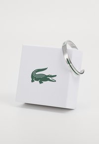 Ezüst színű fémből készült karkötő zöld díszítő csíkokkal, egy fehér dobozon bemutatva, amelyen zöld krokodil logó található. Sima, fényes felület.