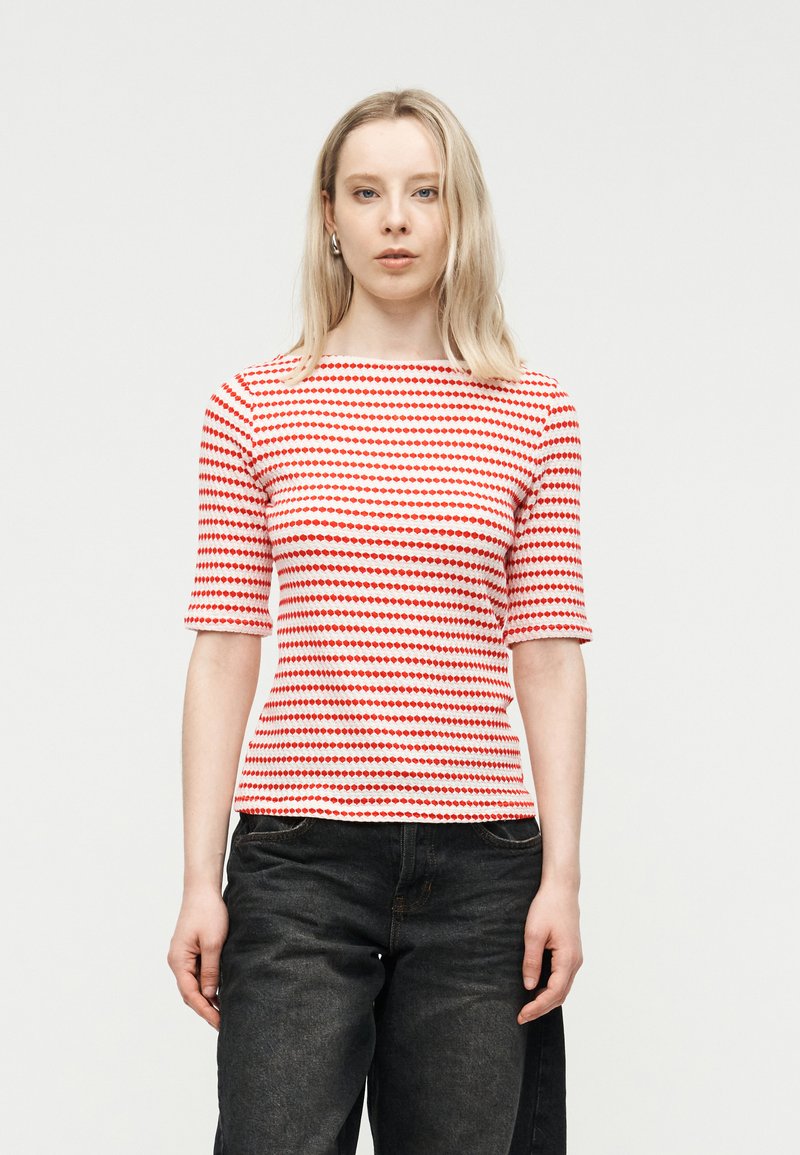 Νεαρή γυναίκα που στέκεται φορώντας ένα κοντομάνικο μπλουζάκι με οριζόντιες κόκκινες και λευκές ρίγες και μαύρο τζιν, μπροστά σε απλό λευκό φόντο.