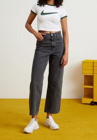 Valkoinen lyhennetyllä mitalla varustettu t-paita, jossa vihreät yksityiskohdat ja Nike-logo, yhdistetty korkeavyötäröisiin, tummanharmaisiin leveälahkeisiin farkkuihin ja valkoisiin lenkkareihin.