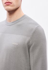 Pulover gri cu textură lăcuită pe umeri, guler rotund și mic logo „BOSS” brodat pe piept. Material neted. Vedere de aproape.