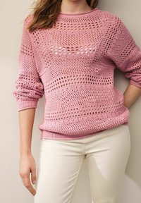 Frau in einem langärmeligen, rosa gehäkelten Pullover mit einem weißen Unterhemd und cremefarbenen Hosen, steht mit einer Hand auf der Hüfte und der anderen nach unten.