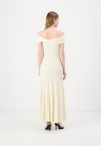 Haut crème ajusté à décolleté off-shoulder se transforme en une jupe longue plissée de couleur jaune doux. La texture est lisse, avec une silhouette fluide. Talons hauts noirs.