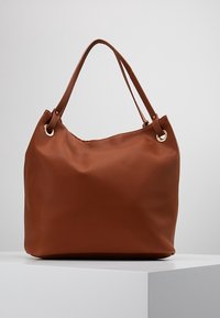 Brun læderhåndtaske med en tekstureret overflade, dobbelte håndtag og metalnitter. Designet er blødt og afslappet med et rummeligt indre.