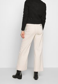 Giacca nera in suede con design aderente, abbinata a pantaloni beige chiaro a gamba larga. Stivali neri alla caviglia con superficie texturizzata e tacco a blocco.