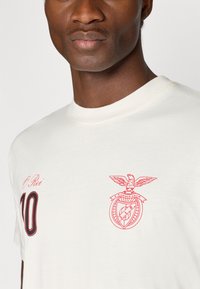 Κρεμ t-shirt από βαμβάκι με κόκκινες εκτυπωμένες γραφικές παραστάσεις: η επιγραφή «O Rei» και ένα έμβλημα που απεικονίζει πουλί με κυκλική ασπίδα. Κλασικό λαιμόκοψη.