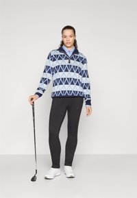 Strikket genser med geometrisk blå og marineblå mønster, glidelås i halsen, svarte leggings og hvite golf sko, holder en golfkølle.