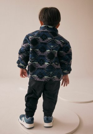 Fleecejacke mit mehrfarbigem geometrischem Muster in Blau- und Lilatönen; schwarze Cargo-Hosen; blau-weiße Sneakers.