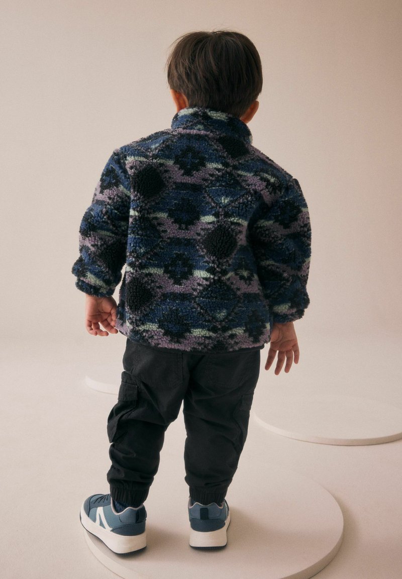 Veste en polaire avec un motif géométrique multicolore dans des tons de bleu et de violet ; pantalon cargo noir ; baskets bleues et blanches.