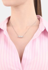 Silverhalsband med tre cirkulära medaljonger; två är vita, en har klara kristaller. Buren över en ljusrosa randig skjorta.