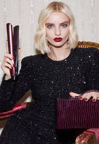 Haarglätter in Roségold und Schwarz mit strukturiertem Griff, gehalten neben einer tiefvioletten Handtasche mit Krokodilprägung, vor einem funkelnden schwarzen Kleid.