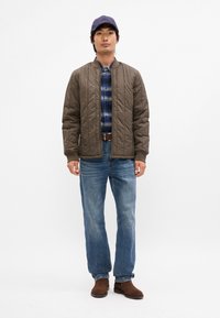 Jachetă maro matlasată, cămașă albastră cu carouri, blugi blue, bocanci maro și o șapcă închisă. Forme și culori simple, textură netedă pe jachetă.