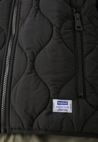 Zwarte gewatteerde vest met een uitgesproken golvend ontwerp, een centrale rits en een label met "HUGO" in het blauw. Textuurstoffen met een zachte afwerking.