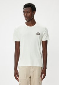 Hvid kortærmet t-shirt med ribbet tekstur, der har et lille sort "NEW YORK" grafik på brystet. Bæres med lyse beige bukser.