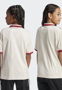 Jersey crème avec un col à rayures rouges et blanches, manches courtes et texture géométrique subtile. Logo en doré près de l'encolure arrière.
