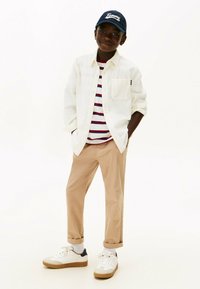 Niño con gorra azul marino, camisa blanca sobre camiseta de rayas, pantalones beiges y zapatillas blancas, de pie con las manos en los bolsillos sobre un fondo blanco.