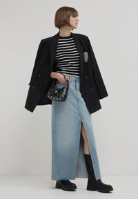 Blazer noir croisé sur une chemise à rayures noires et blanches, jupe en denim clair avec une fente à l'avant, bottines noires, petit sac noir.