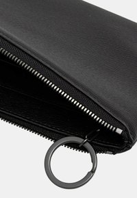 Portefeuille zippé noir en matériau texturé, avec quincaillerie métallique et un anneau d'attache circulaire. L'intérieur est lisse et doublé.