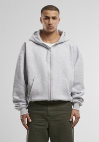 Jeune homme face à l'avant portant un sweat à capuche zippé gris clair et un pantalon vert foncé sur un fond gris uni.