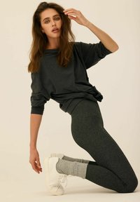 Ciemnoszara, luźna bluza z podwiniętymi rękawami, w połączeniu z ciemnymi legginsami. Modelka ma na sobie białe sneakersy i jasnoszare skarpetki, pozując w pozycji klęczącej.
