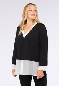 Žena v černém svetr s výstřihem do V, který má navlečený přes bílou košili s knoflíky, stojí na jednobarevném světlém pozadí, dívá se do strany a mírně se usmívá.