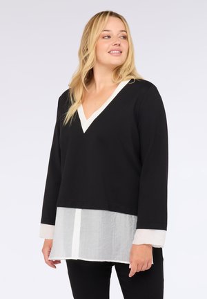 Vrouw in een zwarte V-halstrui, gedragen over een witte blouse met knopen, staand tegen een lichte effen achtergrond, kijkt naar de zijkant en glimlacht lichtjes.