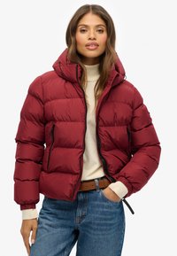 Giacca in piumino rossa con design trapuntato, davanti con zip e cappuccio. Presenta zip nere, polsini elastici e una silhouette aderente.