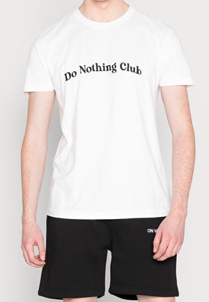 Osoba ubrana w białą koszulkę z napisem "Klub Nicnierobienia" oraz czarne szorty z małym białym napisem na lewym udzie, na tle białym.