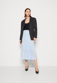 Blazer noir à rayures, haut noir, jupe midi bleu clair avec des couches volantes, et escarpins noirs à bout pointu.