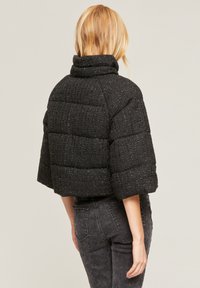 Nainen, jolla on vaaleat hiukset, on pukeutunut mustaan untuvatakkiin, jossa on leveät hihat ja korkea kaulus. Hänellä on tummanharmaat farkut, ja hän seisoo selkä kameraan päin.