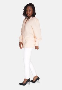 Camicia oversize beige con bottoni, pantaloni bianchi aderenti e décolleté nere a punta, con tessuto testurizzato e maniche arricciate.