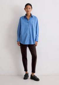 Vrouw in een oversized blauwe overhemd, donkerbruine slim fit broek en zwarte loafers, staand tegen een egale witte muur.
