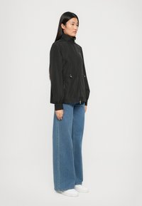 Zwarte lichtgewicht jas met hoge kraag, ritssluiting, elastische manchetten en koord in de taille. Gecombineerd met blauwe jeans met wijde pijpen en witte sneakers.