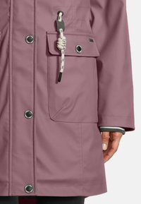 Helle mauve wasserdichte Jacke mit Druckknöpfen, einer großen Fronttasche mit Seil-Detail und einer teilweise sichtbaren Hand an der Seite.